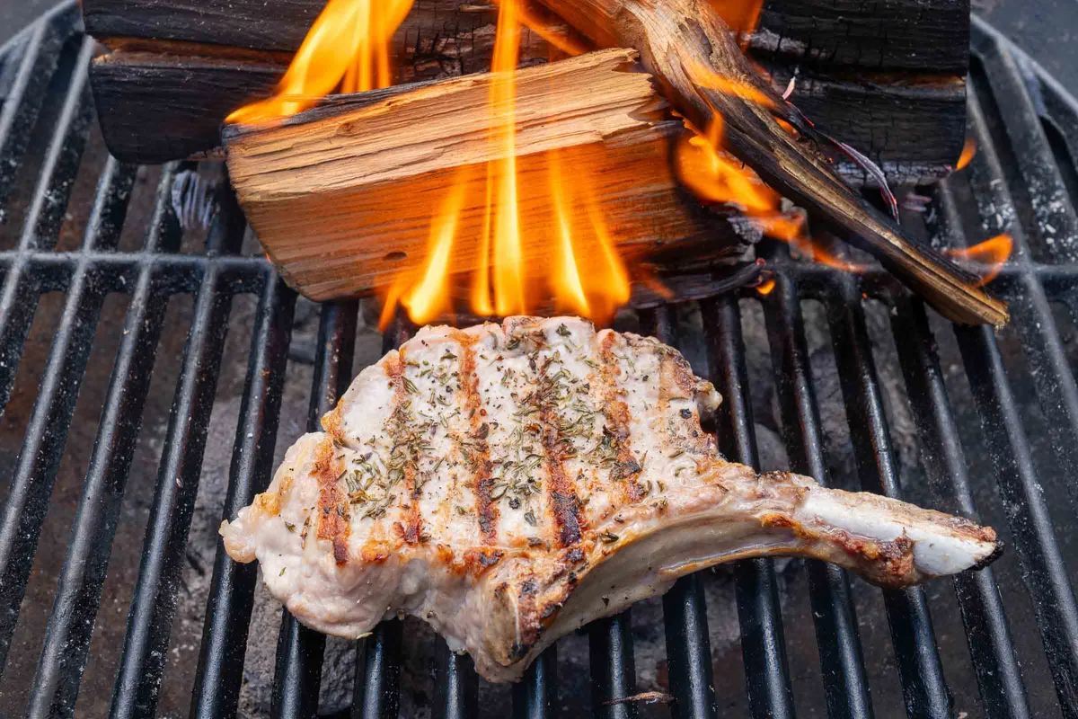 Schweinekotelett am Knochen auf dem Grillrost, dahinter brennende Holzscheite