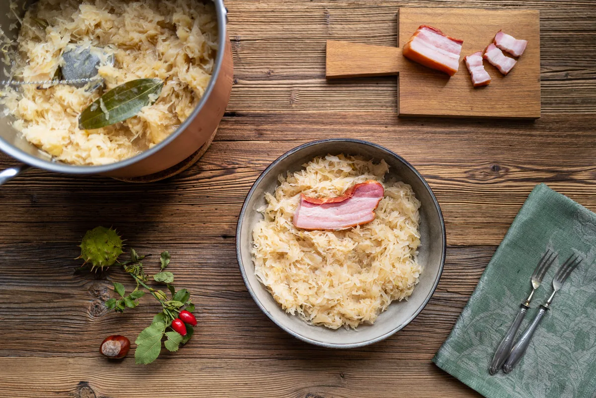 Sauerkraut mit Speckstreifen und Lorbeerblatt in dunkler Schale, Topf im Hintergrund